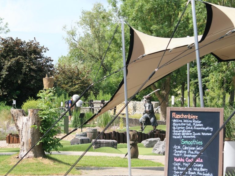 Spielplatz mit Sonnenschutz, Tafel mit Menu, Bäume im Hintergrund und mittelalterliche Statue.